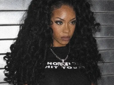 A grainy portrait of a woman with long, voluminous curly black hair wearing a black graphic T-shirt and silver chain necklace, standing in front of a metallic shutter with a serious expression and vintage film-style texture