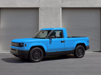 Blue Slate electric pickup photographed from the front three-quarter angle outside gray industrial garage doors