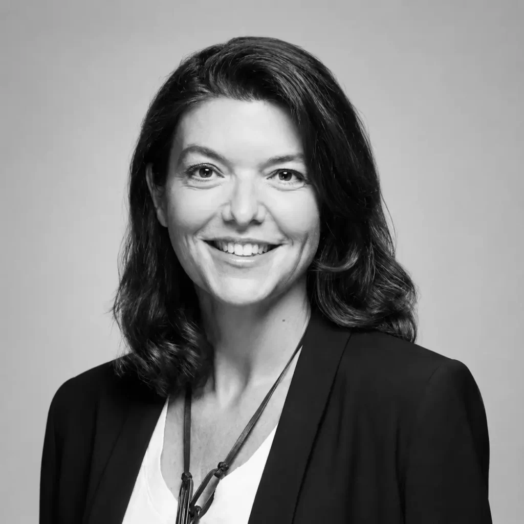 Hélène de Tissot with shoulder-length dark hair, wearing a black blazer over a white top and a long pendant necklace against a dark gray studio background