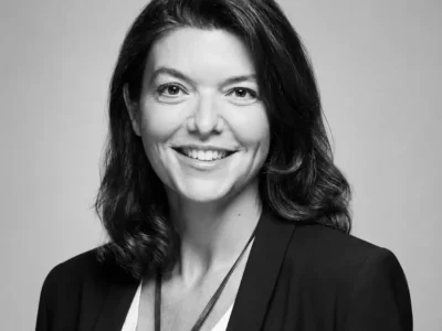 Hélène de Tissot with shoulder-length dark hair, wearing a black blazer over a white top and a long pendant necklace against a dark gray studio background