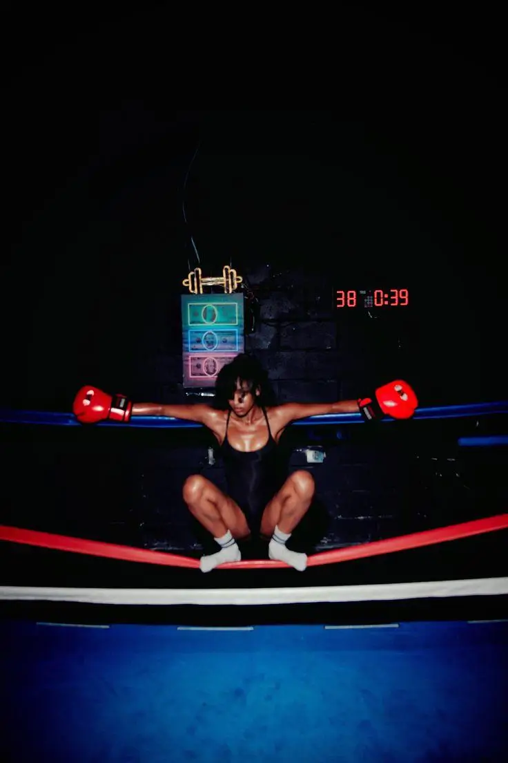 Boxer wearing bright red gloves balances between the ropes of a dimly lit boxing ring beneath neon lights, creating a dramatic training portrait.
