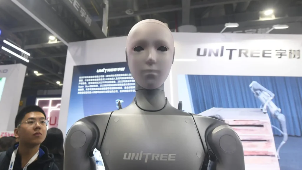 Humanoid robot displayed at the Unitree exhibition booth, featuring a smooth gray face, minimalist facial features, and a metallic body. The trade show environment includes branding, informational displays, and attendees in the background