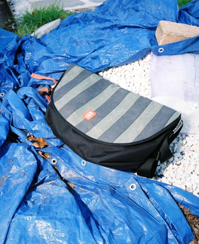 Black and gray striped Crumpler messenger bag placed on blue construction tarps and gravel outdoors, highlighting the rugged materials and reflective striped flap