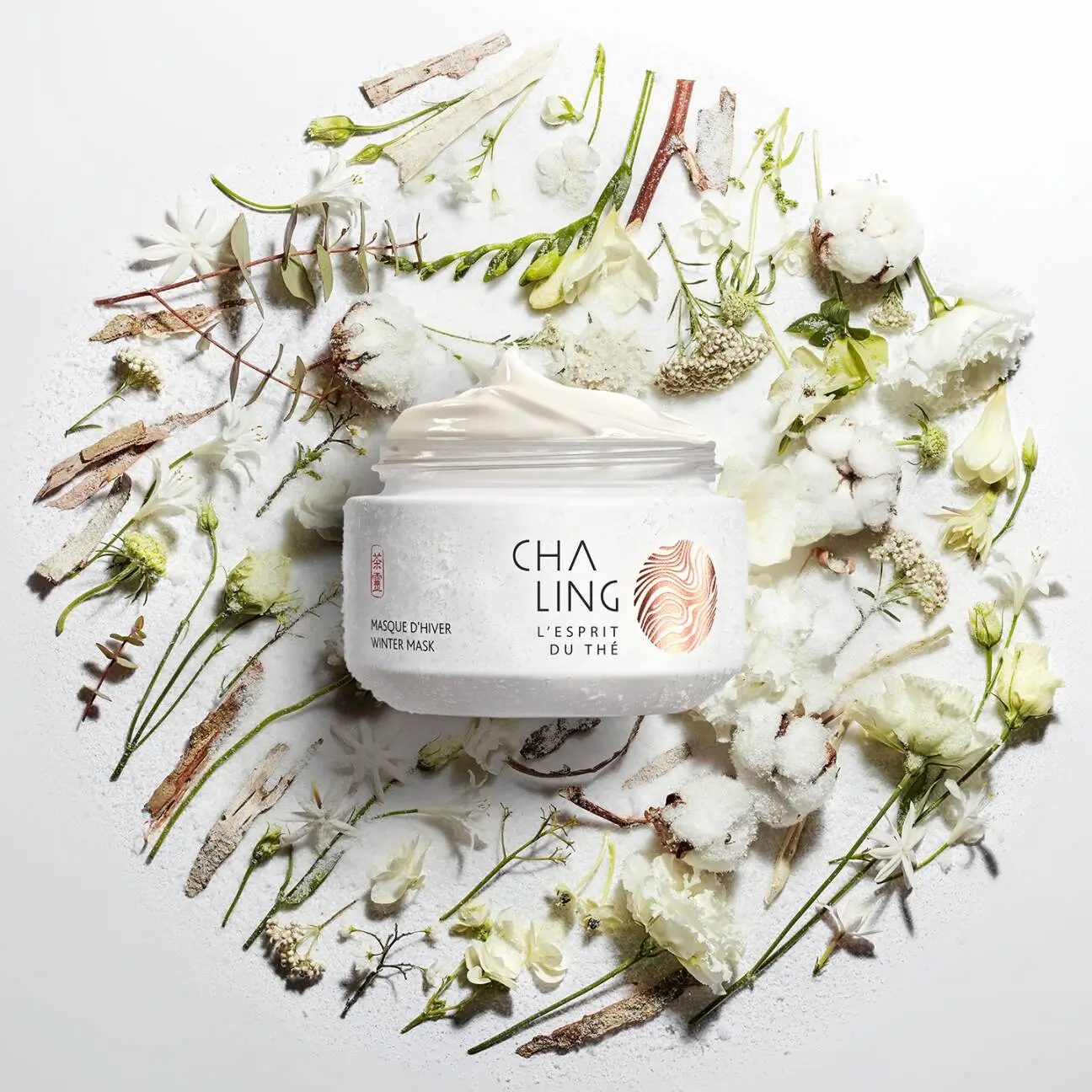 Open jar of CHA LING L'Esprit du Thé Winter Mask skincare cream surrounded by white flowers, cotton bolls, botanical stems, bark, and herbal ingredients arranged on a white background to evoke a clean, nature-inspired luxury beauty composition.
