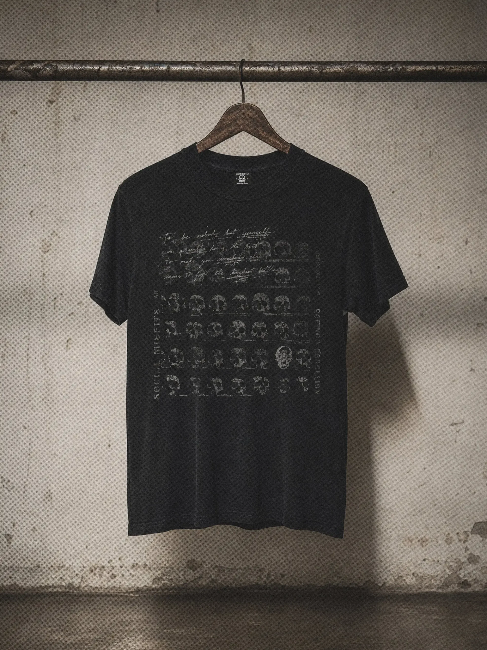 Black graphic T-shirt hanging from a wooden hanger against an industrial concrete wall, featuring a distressed collage of skull illustrations, handwritten script, and faded typography with a vintage washed finish.