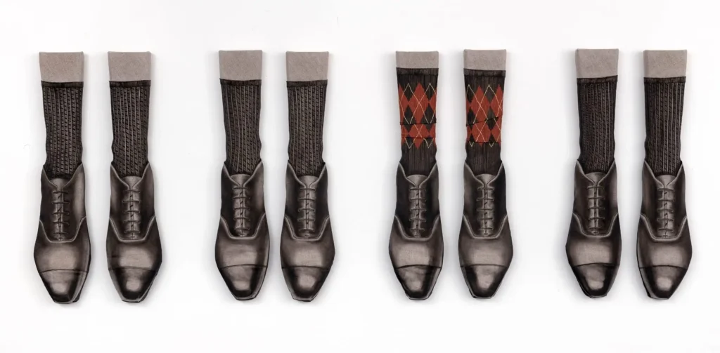 Four pairs of polished black cap-toe dress shoes arranged with tall socks, including ribbed dark socks and a central pair featuring a brown-and-red argyle pattern, presented as a symmetrical textile composition
