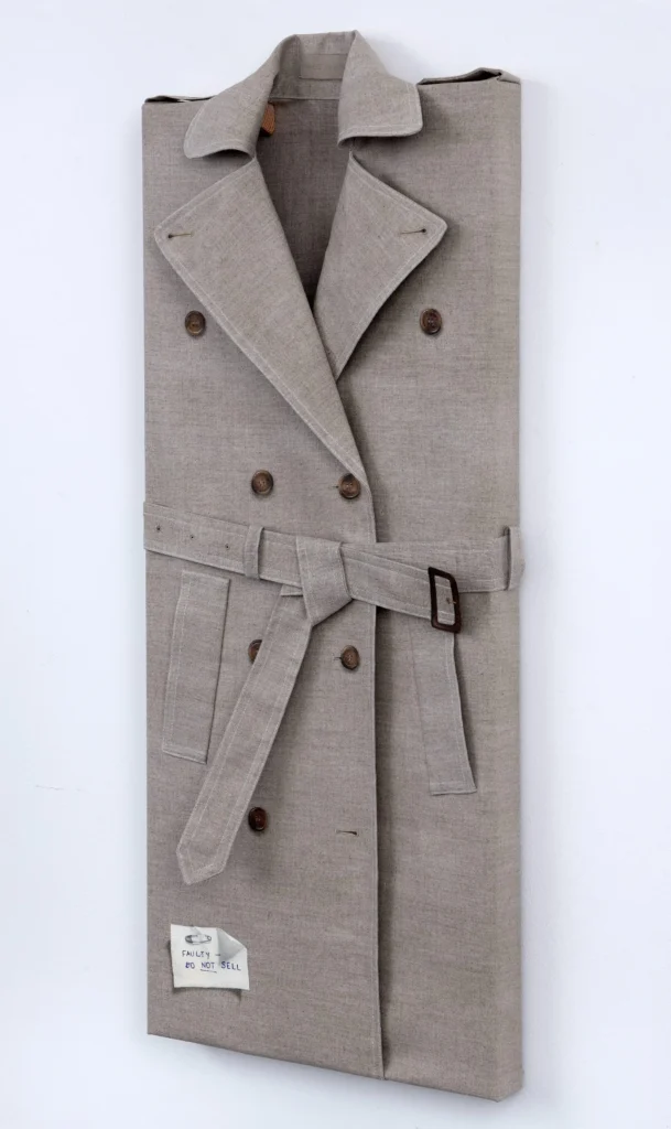 Life-sized canvas artwork of a neatly folded beige double-breasted trench coat with a tied belt, brown buttons, and a handwritten "Faulty – Do Not Sell" tag attached near the hem