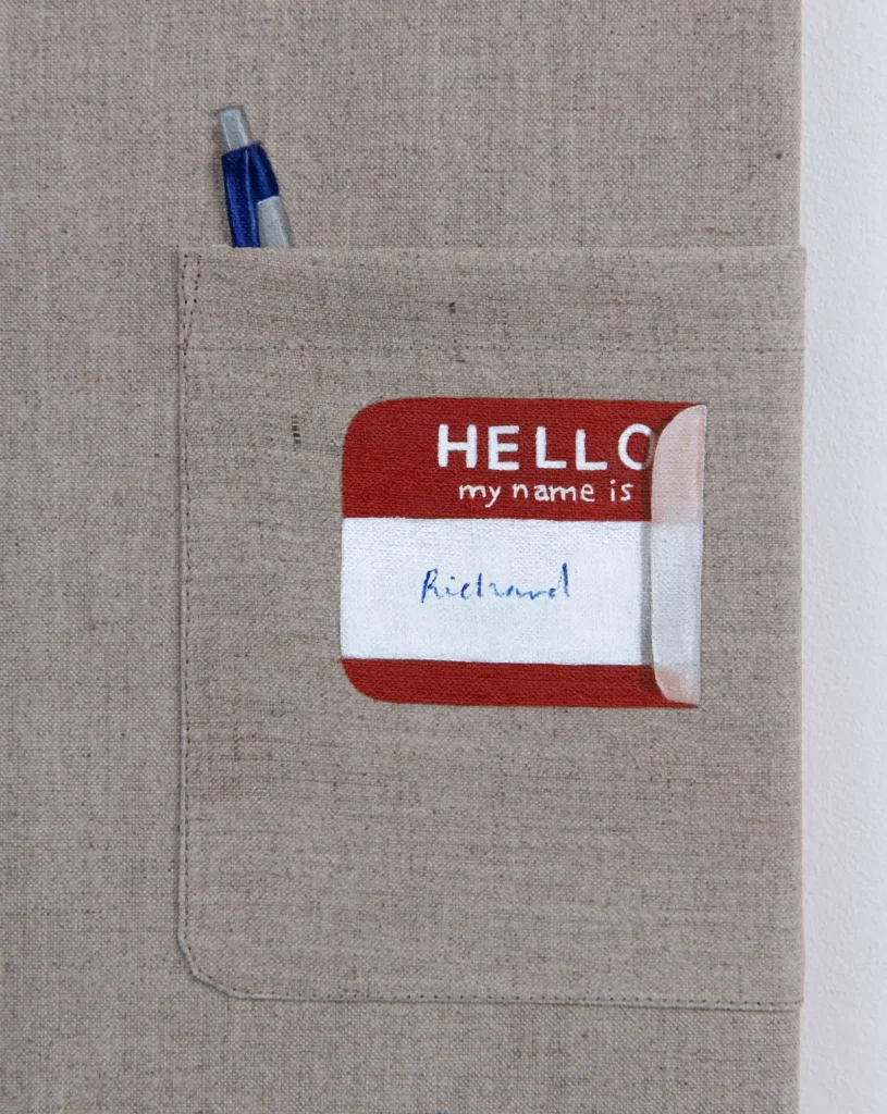 Close-up of a beige fabric shirt pocket featuring a blue pen and a partially peeled red-and-white "Hello My Name Is" sticker handwritten with the name "Richard."