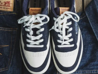 Top-down view of the Reebok x TCB Jeans collide shoe resting on raw indigo denim jeans. The shoes combine white leather toe boxes and side panels with deep indigo denim overlays, white laces, tan leather tongue labels, and co-branded TCB Jeans patches displayed beside the footwear