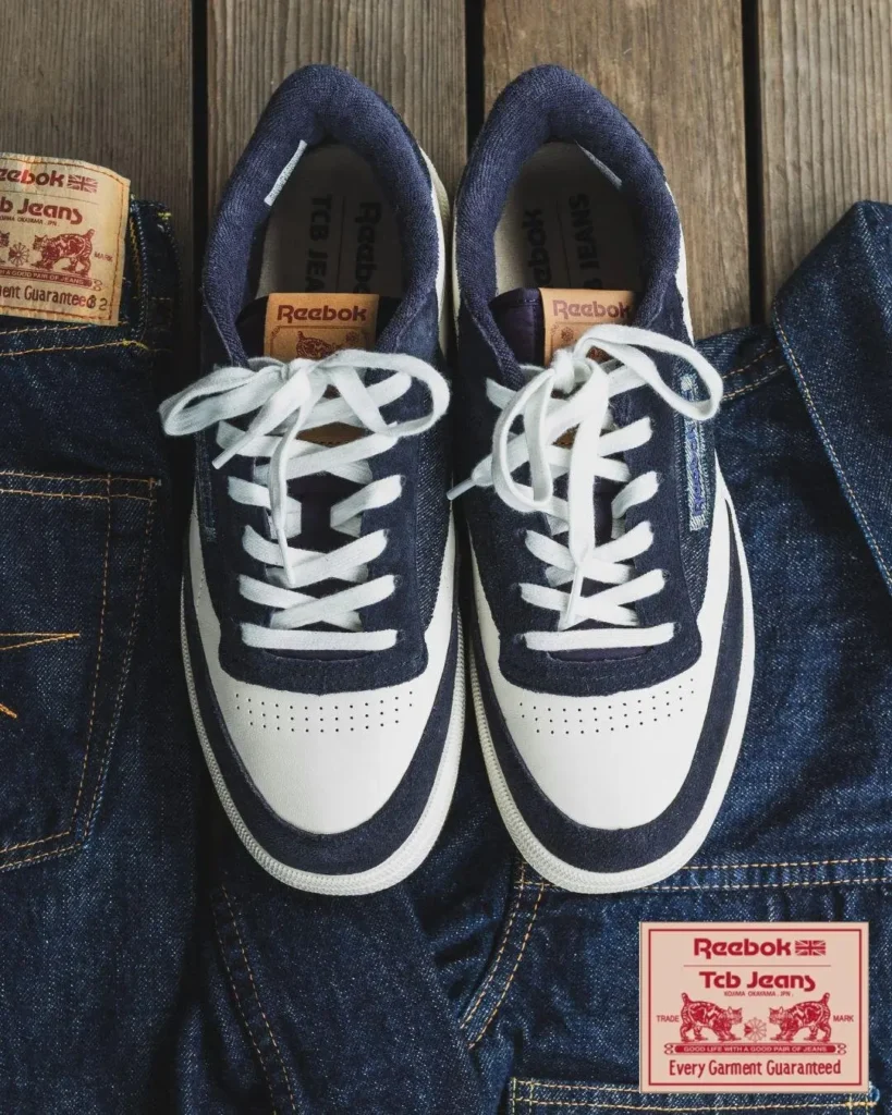 Top-down view of the Reebok x TCB Jeans collide shoe resting on raw indigo denim jeans. The shoes combine white leather toe boxes and side panels with deep indigo denim overlays, white laces, tan leather tongue labels, and co-branded TCB Jeans patches displayed beside the footwear