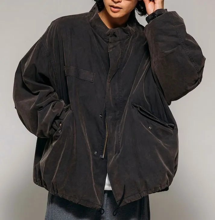 Model wearing an oversized charcoal military-inspired field parka featuring a stand collar, oversized flap pockets, concealed zip and snap front closure, dropped shoulders, adjustable drawcord hem, and a relaxed utilitarian silhouette.