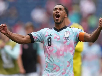 Belgium national team captain celebrates with both fists raised while wearing a pastel blue and pink adidas jersey with a black number 8 and orange captain's armband during a football match
