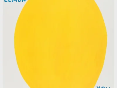 A minimalist poster on a white background featuring an oversized, hand-painted yellow lemon centered on the page with a small green stem. In blue, handwritten uppercase text at the upper left reads, “WHEN LIFE GIVES YOU A LEMON.” At the lower right, matching blue text continues, “YOU MUST EAT THE LEMON,” followed by smaller text beneath that says, “ALL OF IT INCLUDING THE SKIN.” The play illustration uses flat colors and a naïve, graphic style with generous white space surrounding the large fruit