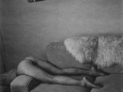 Black-and-white artistic photograph of a person reclining across a fabric sofa, with only their legs visible in black fishnet tights while the rest of their body is concealed behind a large fluffy throw pillow. A chrome arc floor lamp hangs overhead, reflecting the room, and the softly lit, grainy composition creates a moody, vintage-inspired atmosphere with minimalist interior décor