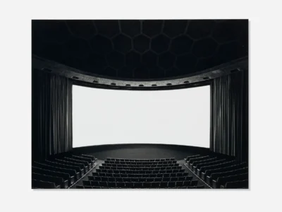 A wide-angle view of an empty modern movie theater with rows of black seats facing a large curved white projection screen. Dark curtains frame the screen, while a geometric honeycomb-patterned ceiling arches overhead, creating a dramatic, symmetrical composition in black and white