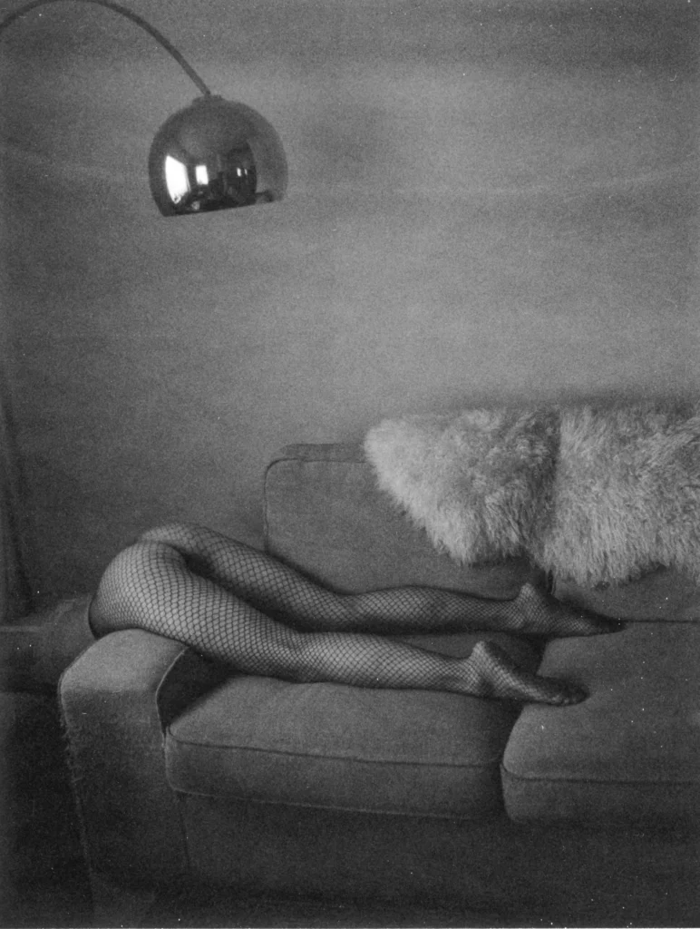 Black-and-white artistic photograph of a person reclining across a fabric sofa, with only their legs visible in black fishnet tights while the rest of their body is concealed behind a large fluffy throw pillow. A chrome arc floor lamp hangs overhead, reflecting the room, and the softly lit, grainy composition creates a moody, vintage-inspired atmosphere with minimalist interior décor