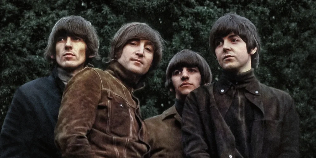 The Beatles pose together outdoors during their mid-1960s era, wearing dark turtlenecks, suede and tailored jackets, and their signature mop-top hairstyles while standing closely against a backdrop of dense green trees