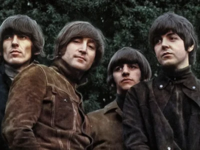 The Beatles pose together outdoors during their mid-1960s era, wearing dark turtlenecks, suede and tailored jackets, and their signature mop-top hairstyles while standing closely against a backdrop of dense green trees