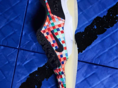 Side profile of the DOAF x Nike shoe displayed on a blue indoor court. The design combines black suede overlays with a vivid woven checkerboard upper, black Swoosh, embroidered DOAF branding on the heel, and a translucent cream outsole