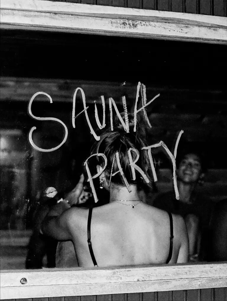 Black-and-white photograph of a person seen from behind writing 'Sunk Party' on a fogged window while people gather inside, creating a candid nightlife scene.