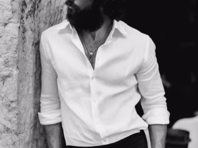 Aaron Taylor-Johnson with curly hair and a full beard wearing dark sunglasses, an open-collar white linen shirt, layered necklaces, dark trousers, and a leather-strap wristwatch, leaning against a textured stone wall in a black-and-white portrait with a relaxed, sophisticated pose