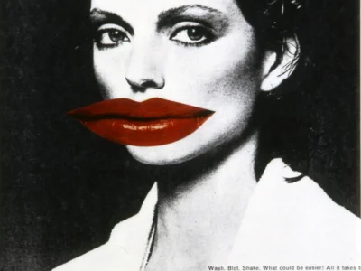 Black-and-white fashion portrait featuring a woman with short dark hair and dramatic eye makeup, overlaid with an oversized pair of glossy red lips across her mouth in a surreal editorial composition. Small magazine text appears in the lower-right corner against the monochrome image