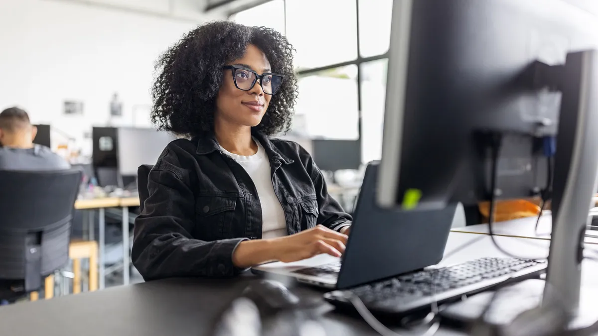 Professional wearing black-framed glasses and a black denim jacket works on a laptop at a desk in a bright open-plan office. A large external monitor, keyboard, and mouse fill the foreground while coworkers and additional workstations are softly blurred in the background.