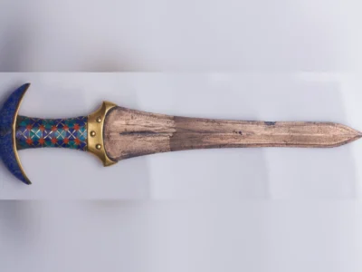 Decorative ancient Egyptian dagger with a weathered bronze blade, gold guard, and a blue crescent-shaped pommel. The handle is adorned with blue, turquoise, and red geometric inlays, displayed horizontally against a plain white background