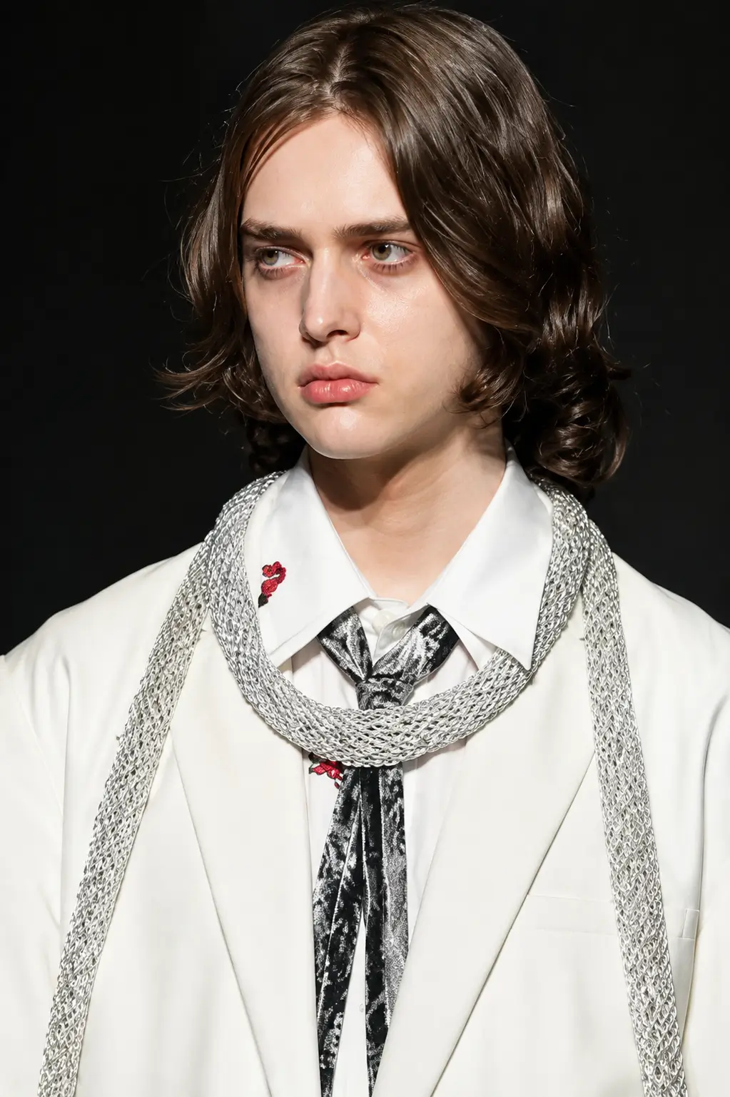 Close-up portrait of a runway model wearing an oversized white suit layered over a white shirt, abstract black-and-silver tie, and thick silver rope necklace with subtle red embroidered accents on the collar.
