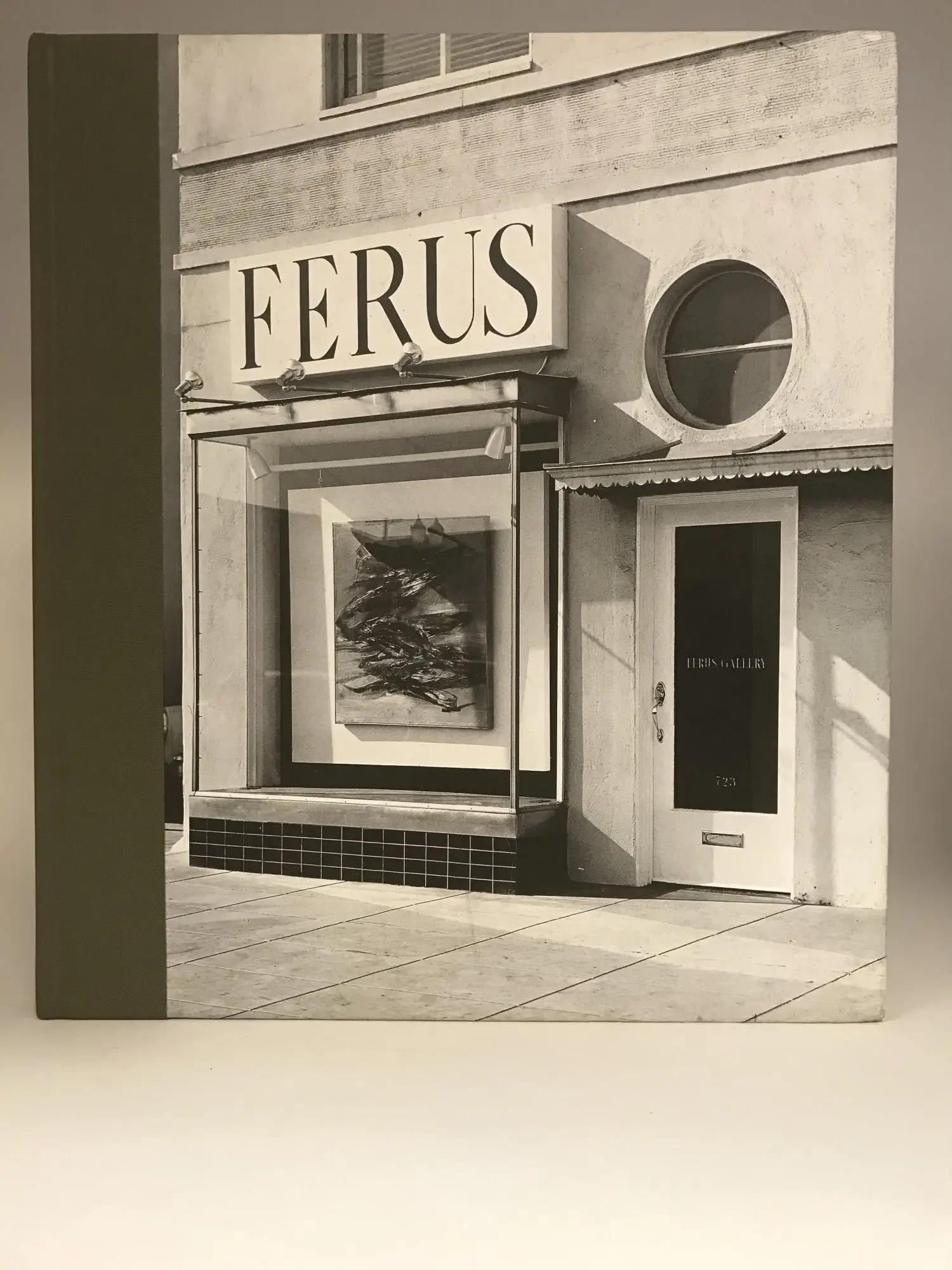 A black-and-white photograph of the historic Ferus Gallery storefront in Los Angeles, featuring its iconic signage above a large display window with an abstract painting inside. The neighboring entrance has a circular window and a door marked 'Ferus Gallery,' capturing the minimalist façade that became a landmark of California's postwar contemporary art scene.