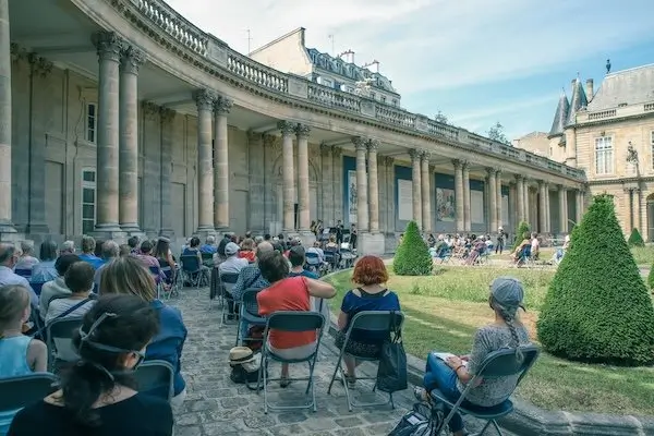 Visitors sit in rows of chairs inside the historic courtyard of Paris's Palais-Royal, attending an outdoor cultural performance framed by elegant colonnades, manicured gardens, and classical French architecture beneath a bright summer sky.