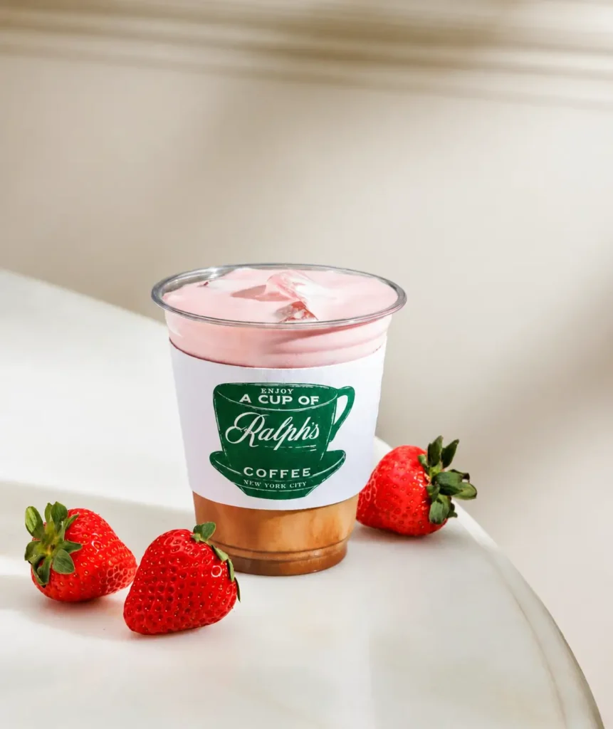 A takeaway cup of Ralph's Coffee strawberry latte rests on a white tabletop, topped with light pink foam and ice beneath a clear lid. The cup displays the green Ralph's Coffee logo sleeve and is surrounded by three ripe strawberries in a bright, minimalist café set
