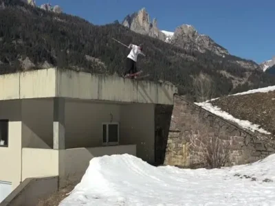 Martin Longhitano slides across the rooftop edge of a modern mountain building before dropping toward a snow-built landing, framed by the rugged peaks of Italy's Dolomites under clear blue skies