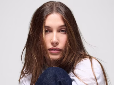 A close-up studio portrait of a model with long chestnut-brown hair parted at the center, gazing directly into the camera with a relaxed expression. She wears a white top while resting against a bent knee, photographed against a clean light gray backdrop with soft, natural light