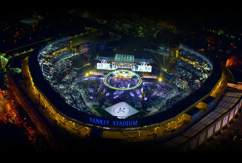 Aerial nighttime view of Yankee Stadium filled with thousands of concertgoers surrounding a large circular stage illuminated by colorful lights, with the stadium exterior and surrounding city streets glowing after dark.