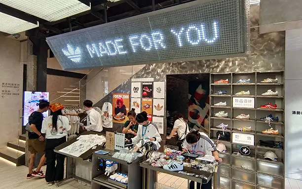 Interior of an adidas retail store featuring a large illuminated 'MADE FOR YOU' sign above a customization counter, with staff assisting customers personalizing sneakers and apparel at work tables, a 'Forum 2000' promotional display, and a wall-mounted shelving unit stocked with rows of adidas sneakers in various colorways.