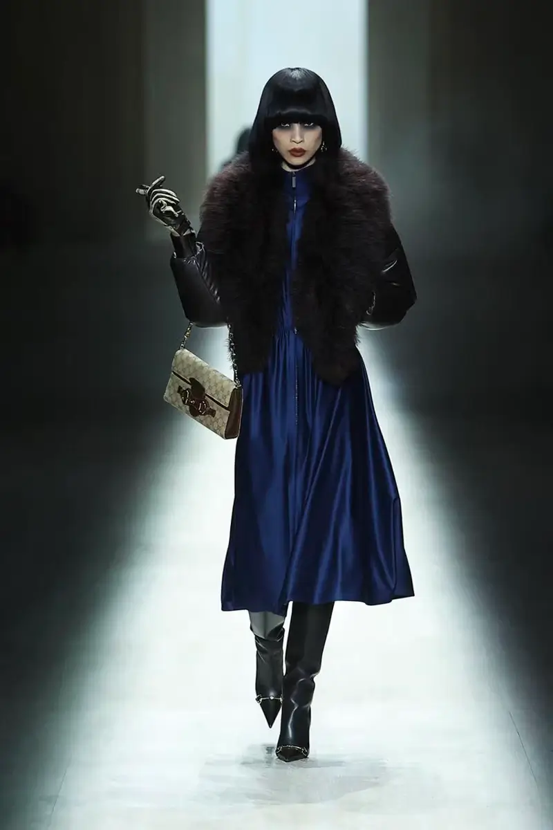 Runway model wearing a deep blue satin midi dress layered with a dark fur stole, black leather gloves, knee-high boots, and a beige monogram shoulder bag during a luxury fashion show.
