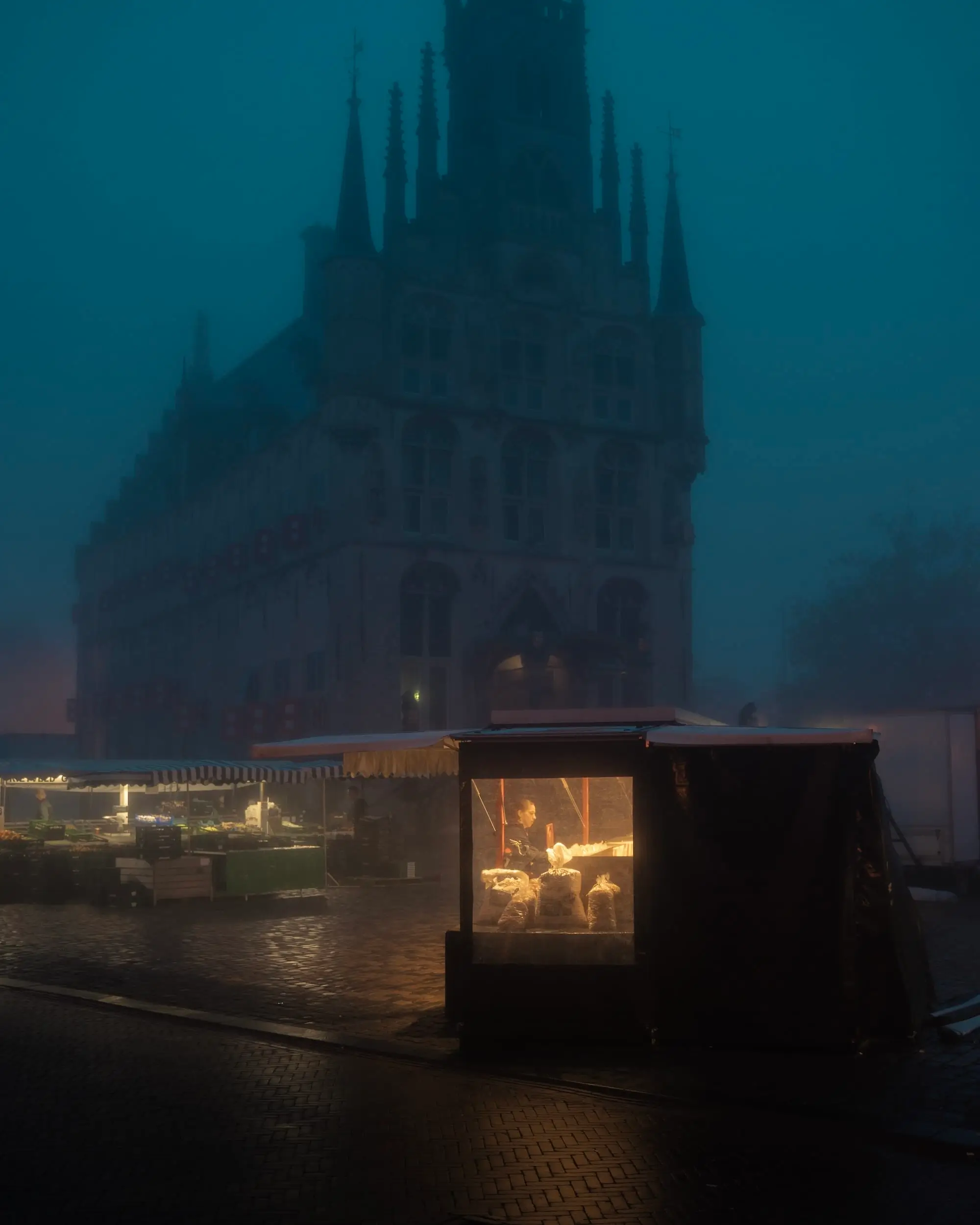 Foggy European market square at dawn with a warmly lit vendor stall in the foreground and a Gothic church rising through the mist in the background.