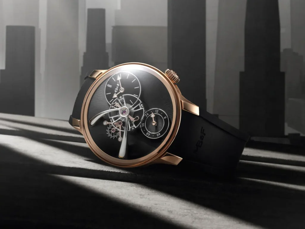 Close-up of the MB&F Legacy Machine 101 EVO resting on a shadowed surface, showcasing the polished rose gold case, black dial, exposed flying balance wheel, offset power reserve and time subdials, with an architecture grayscale backdrop