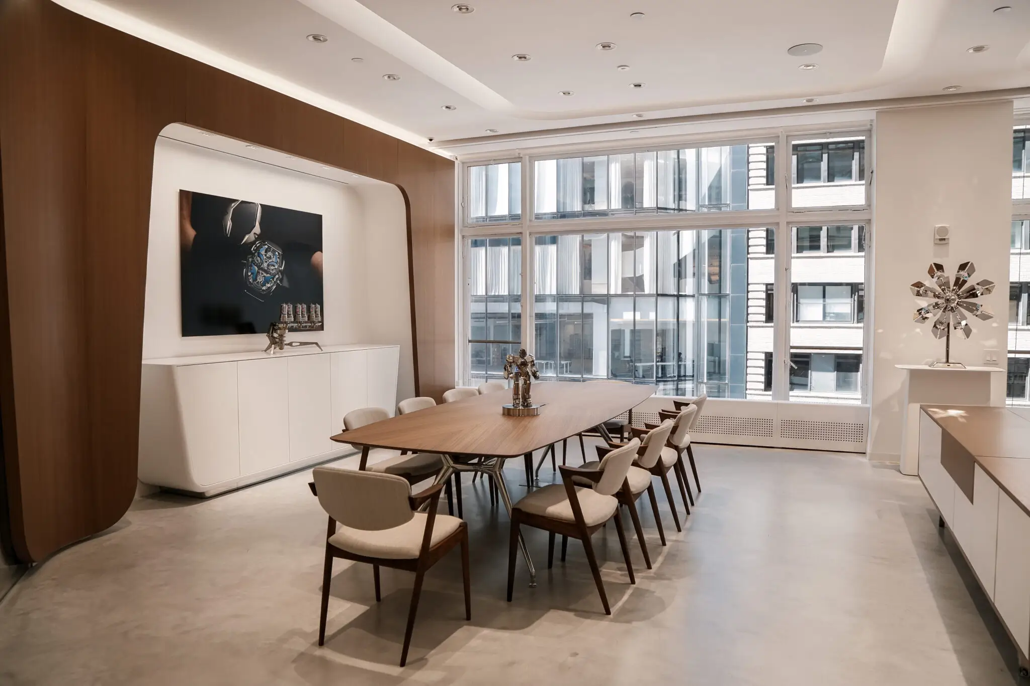 Contemporary MB&F New York lounge with a walnut conference table, upholstered chairs, floor-to-ceiling windows, kinetic sculptures, and a large horology artwork displayed within a warm wood-paneled interior.