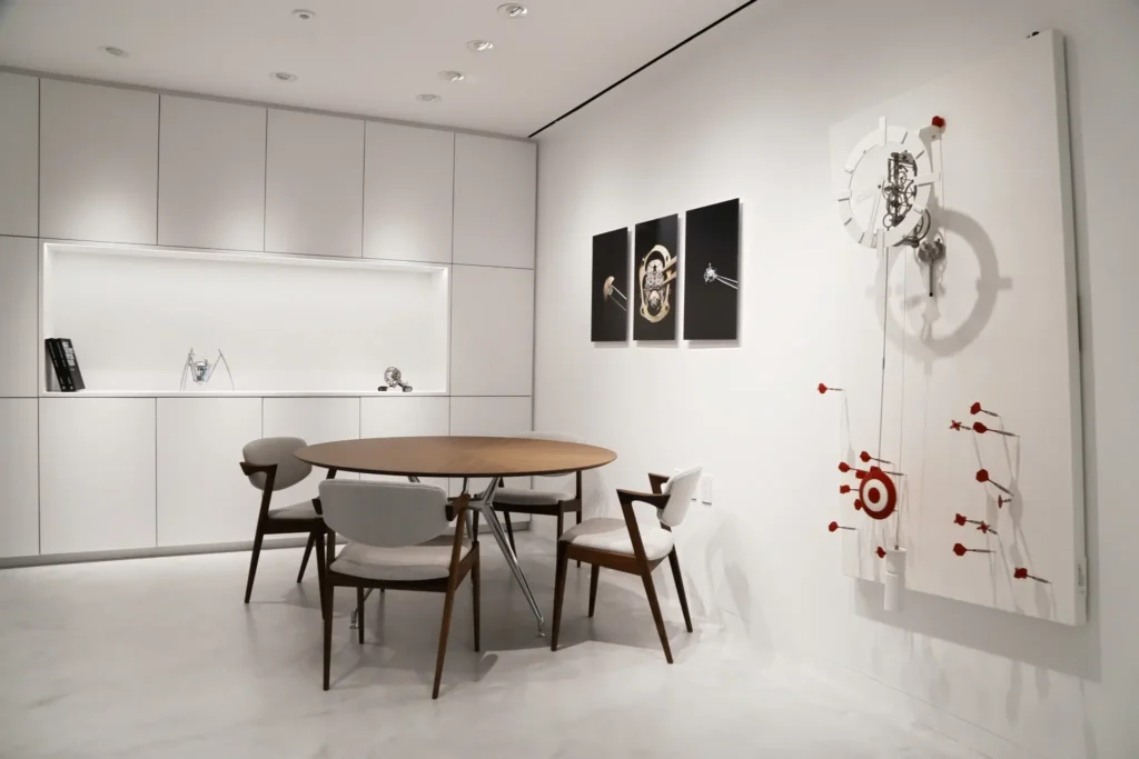 Minimal private meeting room inside the MB&F New York lounge with a round wooden table, modern chairs, built-in illuminated shelving, horological artwork, and a kinetic wall sculpture accented with red element