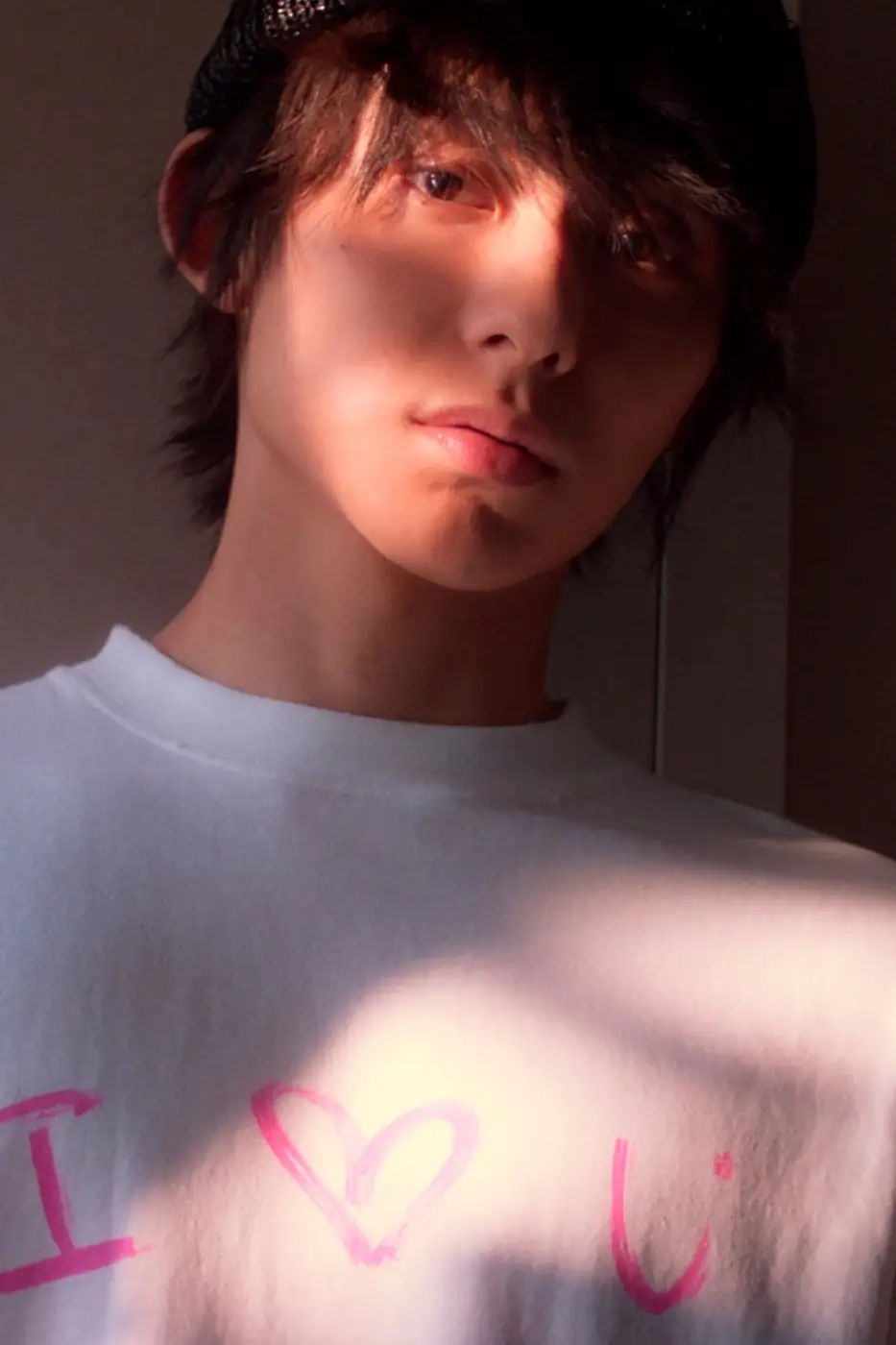 Close-up portrait of a person wearing a black knit beanie and an oversized white Balenciaga T-shirt with a hand-drawn style pink 'I ♥ U' graphic. Warm sunlight casts dramatic bands of light and shadow across the face, creating an intimate mood against a softly darkened interior background.