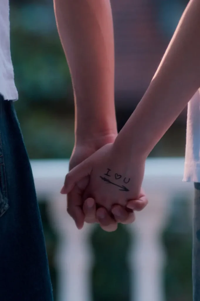 Close-up of two people holding hands outdoors. One hand has "I ♥ U" written in black marker with an arrow pointing toward the other person, emphasizing a playful and affectionate gesture against a softly blurred background