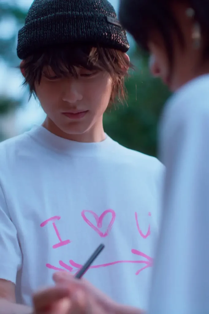A person in a black knit beanie and oversized white T-shirt stands while another person writes on the front of the shirt with a black marker. The shirt displays a bright pink "I ♥ U" graphic with a large arrow beneath it, capturing a candid moment between the pair