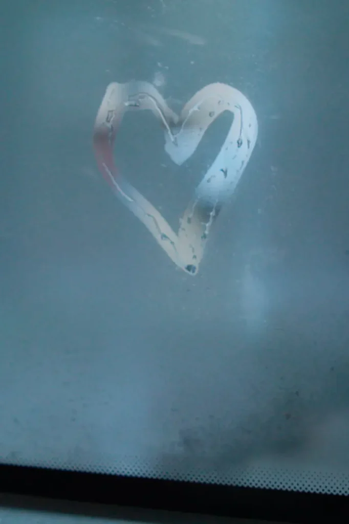 A heart shape traced into a fogged window or glass surface. Water droplets cling to the edges of the hand-drawn heart, creating a simple romantic detail against the cool blue-gray background