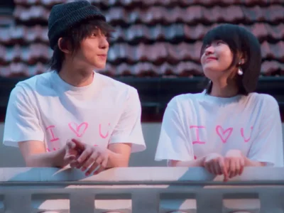 Two people wearing matching oversized white "I ♥ U" graphic T-shirts lean against a white balcony railing at dusk. They exchange warm smiles while facing each other, with tiled rooftops and soft evening light creating a shh, romantic atmosphere