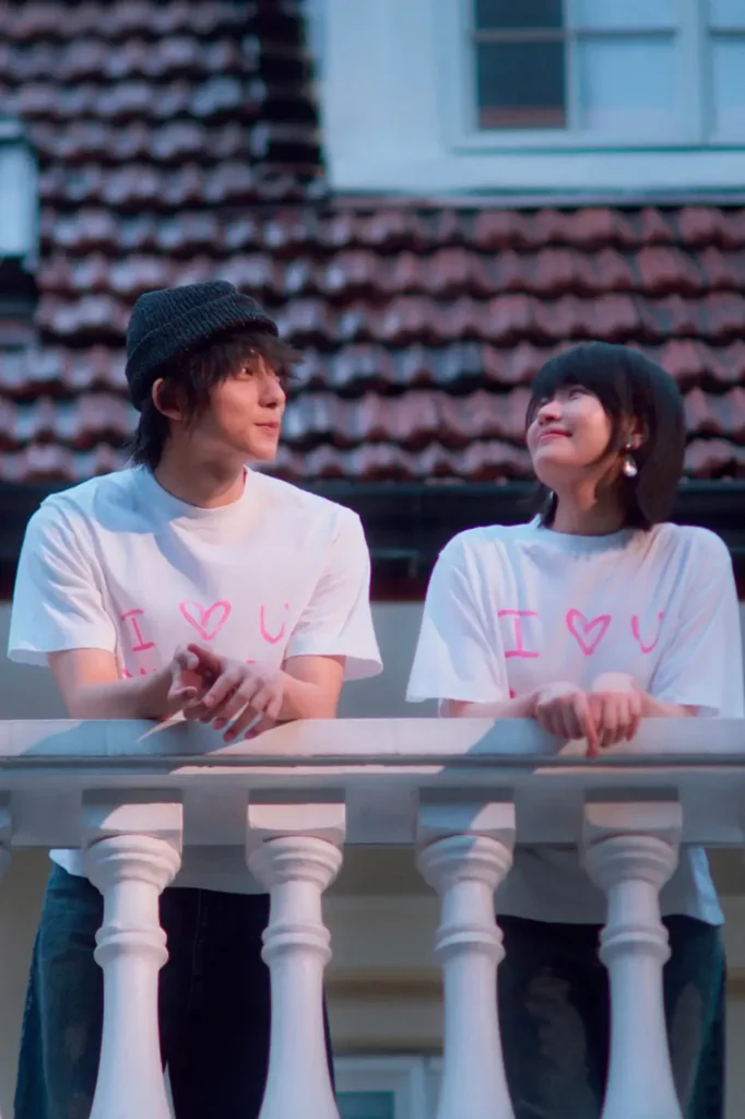 Two people wearing matching oversized white "I ♥ U" graphic T-shirts lean against a white balcony railing at dusk. They exchange warm smiles while facing each other, with tiled rooftops and soft evening light creating a shh, romantic atmosphere