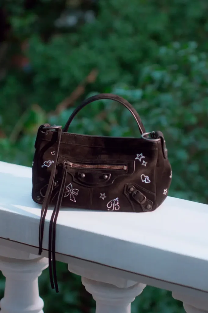 A black Balenciaga shoulder bag rests on a white balcony railing. The bag is decorated with sparkling rhinestone-style doodles, including hearts, bows, stars, and floral motifs, blending the house's signature silhouette with playful, hand-drawn-inspired embellishment