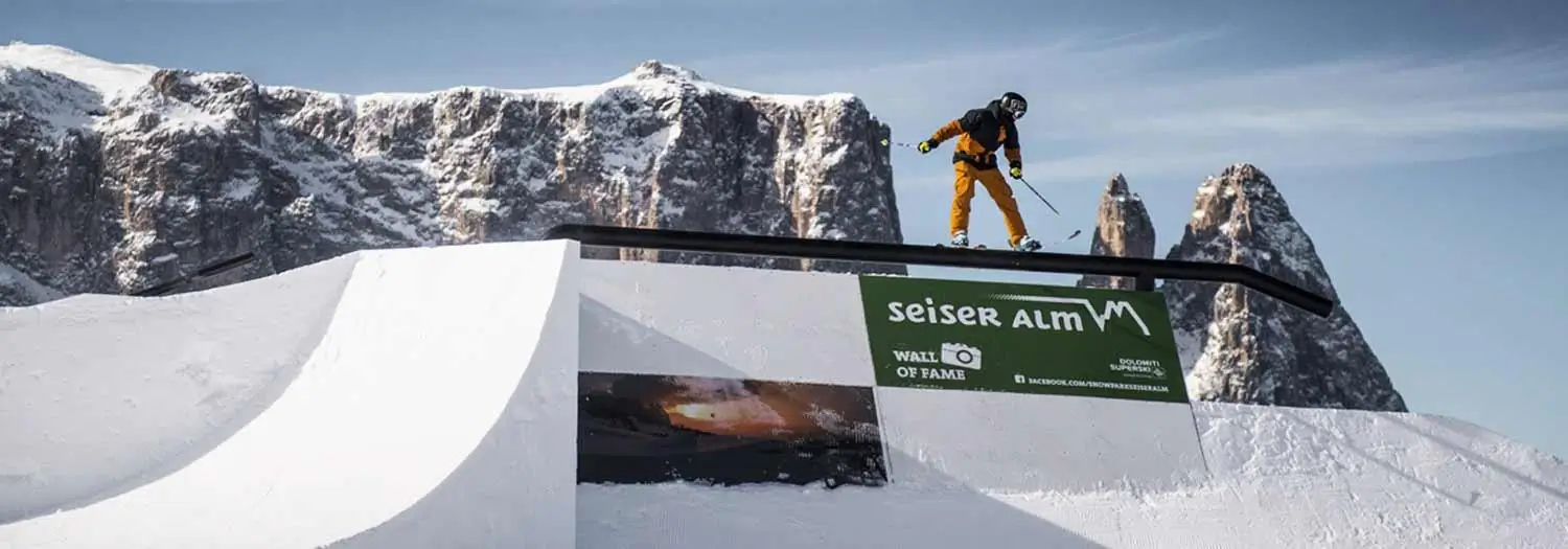 Martin Longhitano rooftop skiing in Italy's Dolomites, balancing across the edge of a modern building before dropping toward a snow-built landing with rugged alpine peaks in the background.