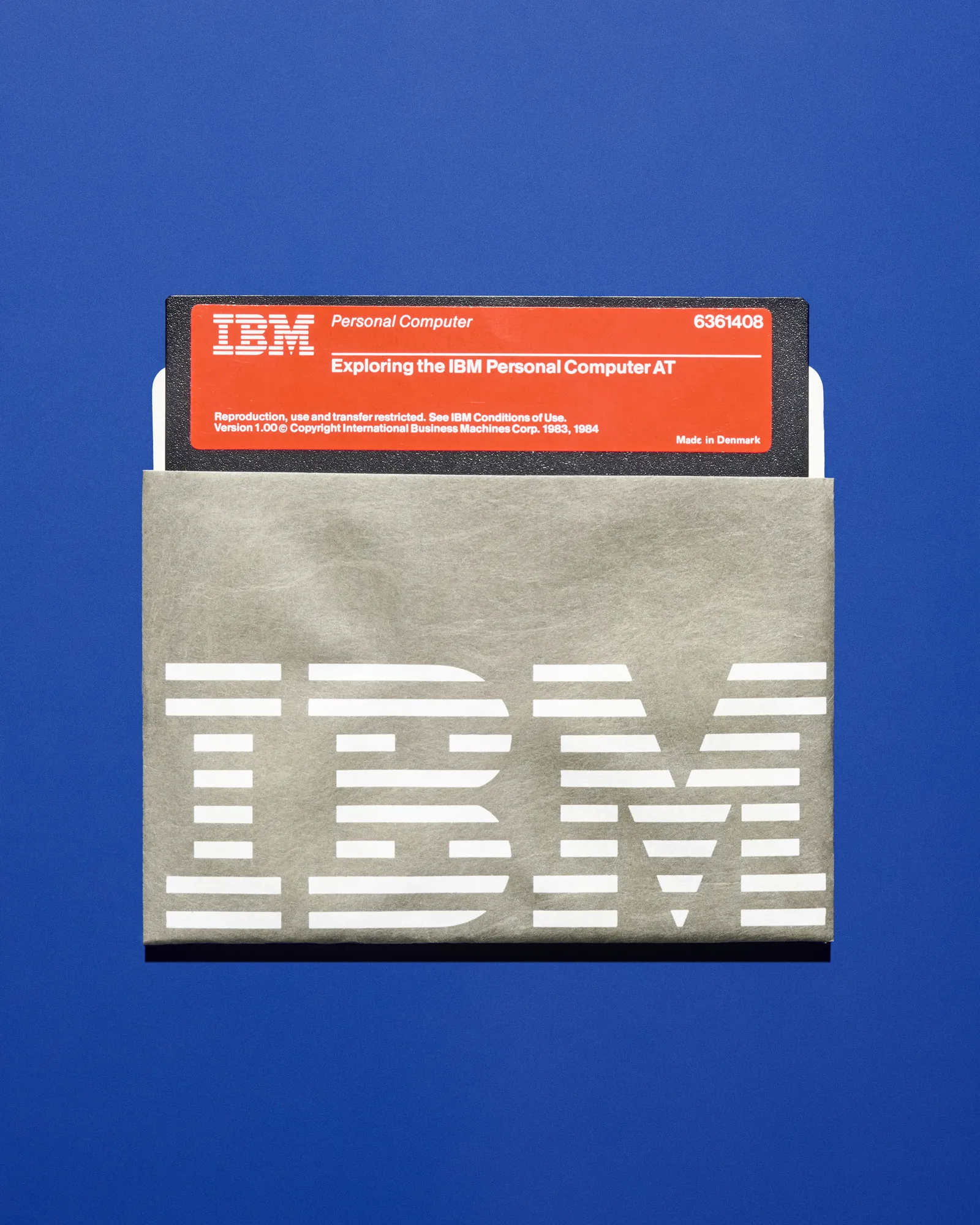 Vintage IBM paper floppy disk sleeve featuring the iconic striped IBM logo with a red 'Exploring the IBM Personal Computer AT' floppy disk partially inserted, photographed against a deep blue background.